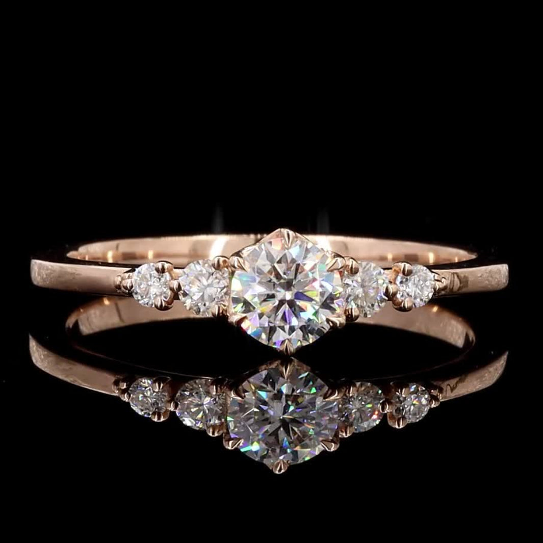 A rose gold diamond ring with a central round brilliant-cut diamond and smaller side diamonds, displayed on a reflective black surface against a black background.