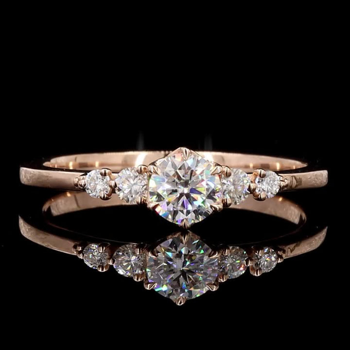 A rose gold diamond ring with a central round brilliant-cut diamond and smaller side diamonds, displayed on a reflective black surface against a black background.