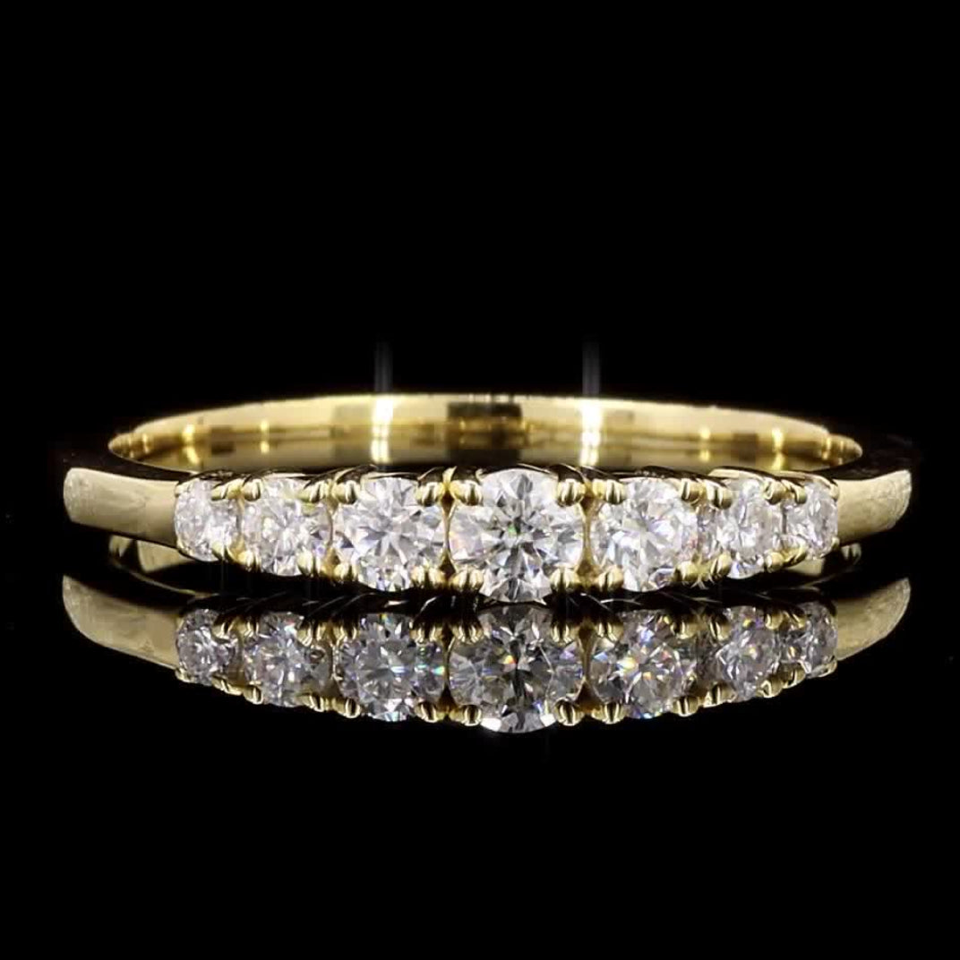 A yellow gold eternity ring featuring a row of round-cut clear gemstones, displayed on a reflective black surface against a solid black background.