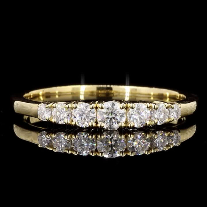 A yellow gold eternity ring featuring a row of round-cut clear gemstones, displayed on a reflective black surface against a solid black background.