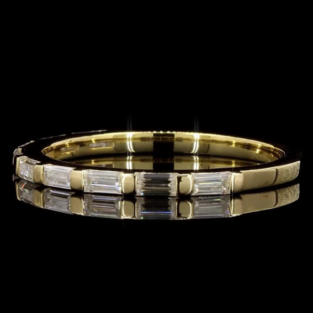 A yellow gold eternity ring featuring baguette-cut clear stones, shown on a reflective black surface against a solid black background.