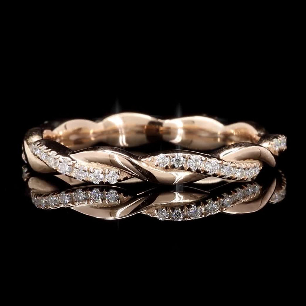 A twisted rose gold ring with pavé-set clear stones, displayed on a reflective black surface against a solid black background.