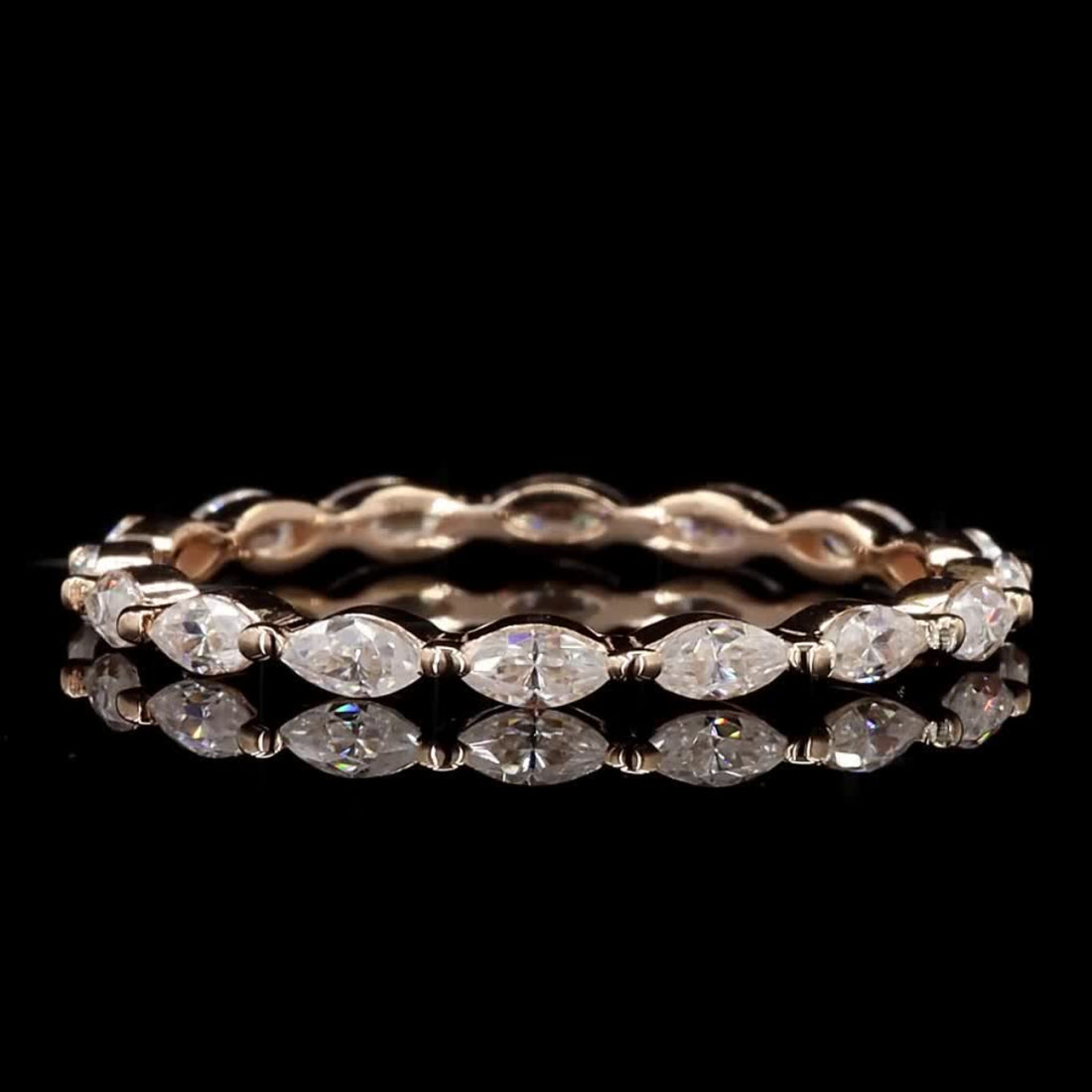 A rose gold eternity ring featuring marquise-cut clear gemstones, displayed on a reflective black surface against a solid black background.