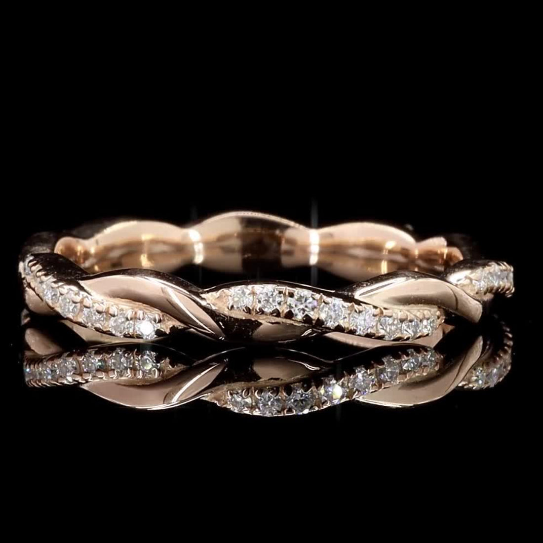 A twisted band ring in rose gold with pavé-set clear stones, photographed on a reflective black surface against a black background.