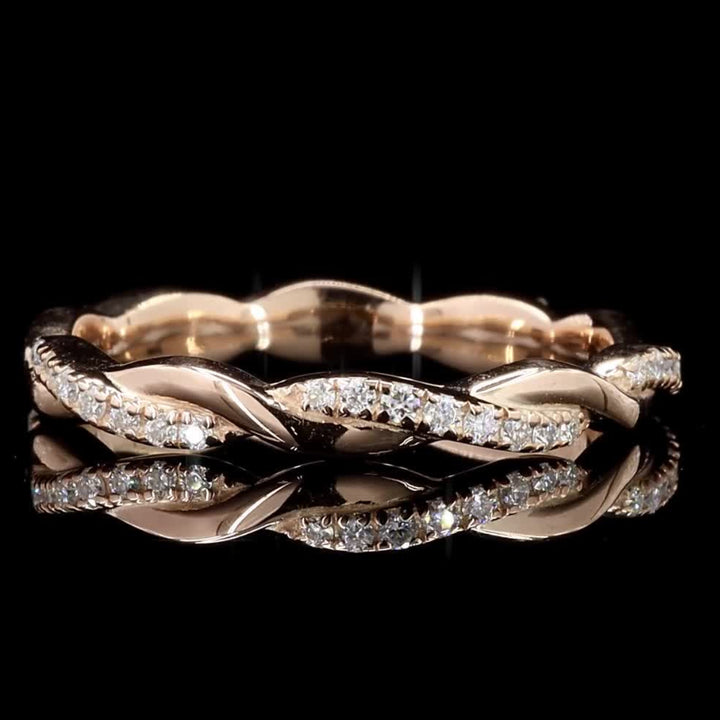 A twisted band ring in rose gold with pavé-set clear stones, photographed on a reflective black surface against a black background.